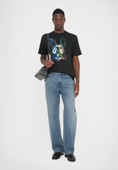 Black T-shirt with a colorful rabbit graphic, paired with loose light blue jeans and black shoes. Bag slung over shoulder, casual fit.