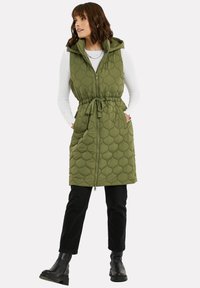 Quilted grüner Weste mit Kapuze, taillierter Taille, zwei seitlichen Taschen und einem Reißverschluss vorne. Kombiniert mit schwarzen Jeans und Stiefeletten.