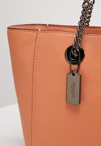 Sac en cuir grainé couleur cognac avec anses chaîne en métal noir et une étiquette métallique noire gravée "Coach New York".