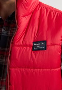 Rote Pufferweste mit Reißverschluss und einem schwarzen Stoffetikett, auf dem "Petrol Ind." steht. Strukturiert, glatte Oberfläche mit sichtbaren Nähdetails.