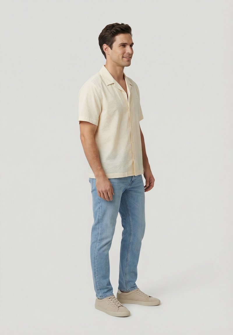 Homme debout, portant une chemise crème à manches courtes boutonnée, un jean bleu clair et des baskets beiges sur un fond clair uni.