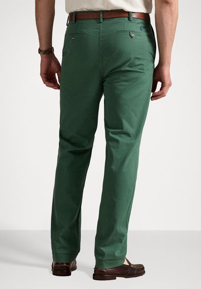 Zaļi kokvilnas chinos bikses ar taisnu kāju, aizmugures kabatām un brūno jostu. Izceļas ar diskrētu logotipu kreisajā pusē. Gluda tekstūra.
