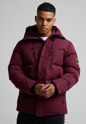 Jeune homme portant une veste d'hiver matelassée bordeaux avec capuche, debout et joignant ses mains devant un fond uni gris clair.