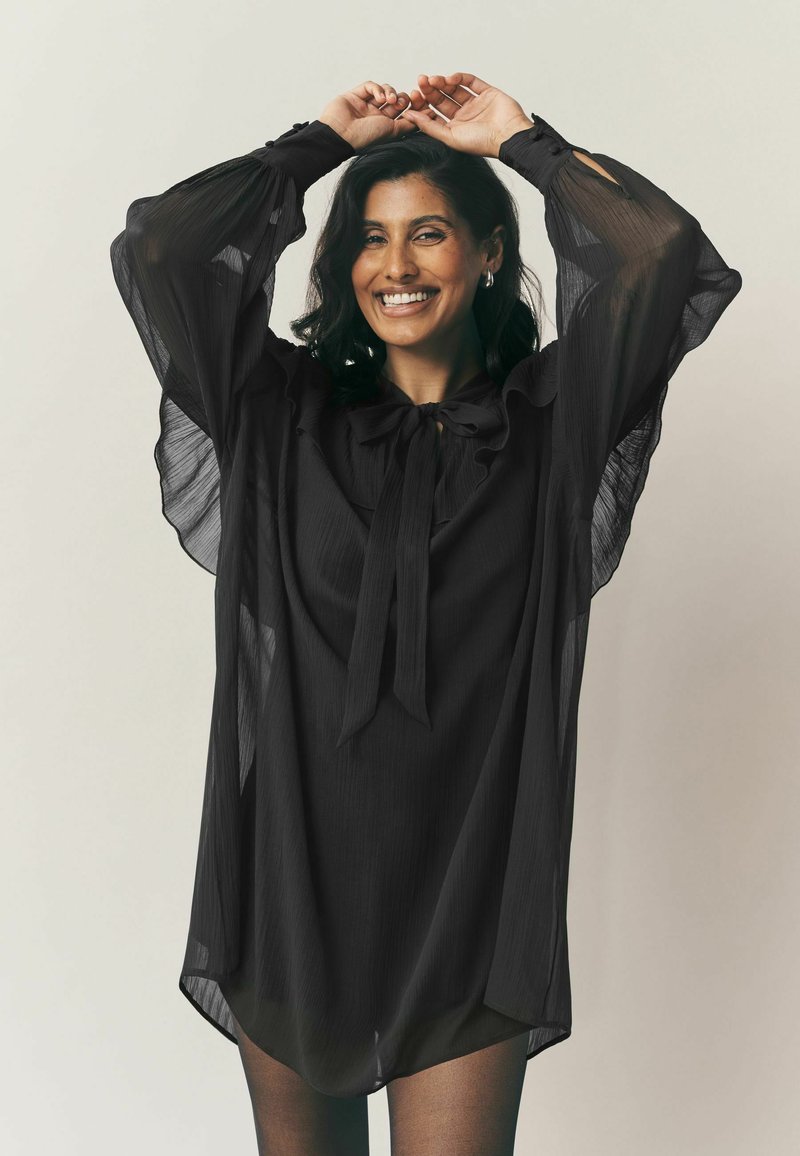 Fekete áttetsző blúz hosszú, puffos ujjakra. Nagy masni a nyakán és laza, folyó sziluett. Sima textúra.