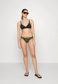 Top de biquíni preto com design triangular, combinado com cuecas de biquíni verde com padrão camuflado. Modelo usando óculos de sol e chinelos.