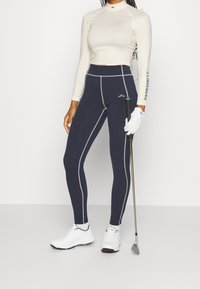 Námořnické sportovní legíny se světle šedými švy, v kombinaci s dlouhým rukávem vzorovaným topem v bílém a zlatém, a bílými golfovými botami. V ruce golfová hůl.