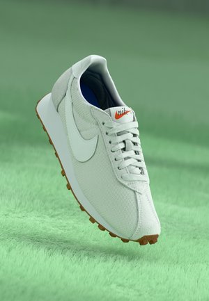 Biały sneaker Nike z detalami z siateczki i zamszu, gumową podeszwą oraz pomarańczowym logo na języku, na zielonym teksturowanym tle.