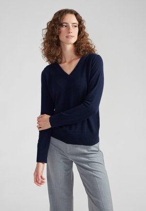 Suéter de cuello en V en color marino, confeccionado en un suave tejido de punto, presenta mangas largas con puños acanalados y un dobladillo acanalado, combinado con pantalones ajustados en gris claro.