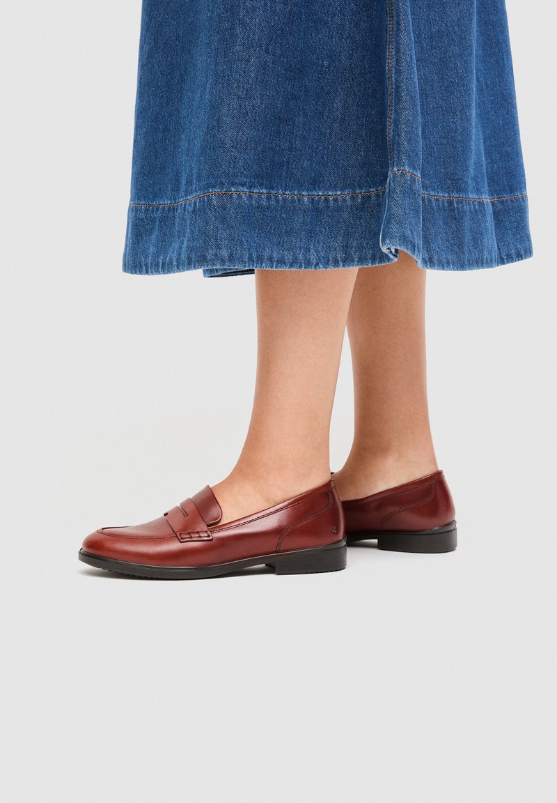 Kvinne som har på seg en blå denimskjørt i middels lengde og brune skinnloafers, står på et lysegrått gulv.