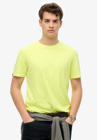 Camiseta de algodón de manga corta y cuello redondo en un brillante color amarillo, combinada con jeans oscuros y un suéter gris atado a la cintura. Detalle notable de reloj de pulsera.
