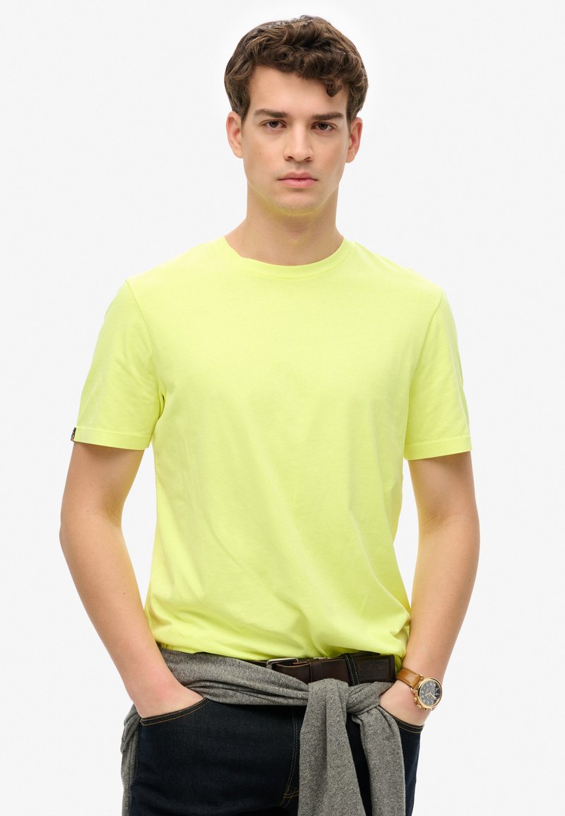 Camiseta de algodón de manga corta y cuello redondo en un brillante color amarillo, combinada con jeans oscuros y un suéter gris atado a la cintura. Detalle notable de reloj de pulsera.