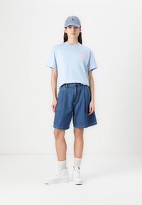 Ljusblå bomullströja med rosa grafik, som kombineras med löst sittande blå jeansshorts. Vita högtoppsskor och matchande keps.