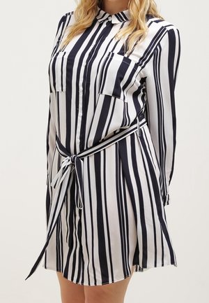 Woman wearing a long-sleeve, black and white vertical striped shirt dress with a collar, chest pockets, and a waist tie belt.