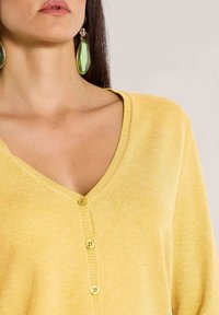 Gelber Cardigan mit V-Ausschnitt, gerippten Abschlüssen und drei gelben Knöpfen auf der Vorderseite. Weiche Textur mit leichtem Erscheinungsbild.