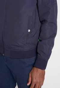 Granatowa kurtka bomberka z gładkiego materiału, z zapięciem na zamek błyskawiczny z przodu, kieszenią na zatrzask, elastycznymi mankietami oraz zieloną metką akcentującą.