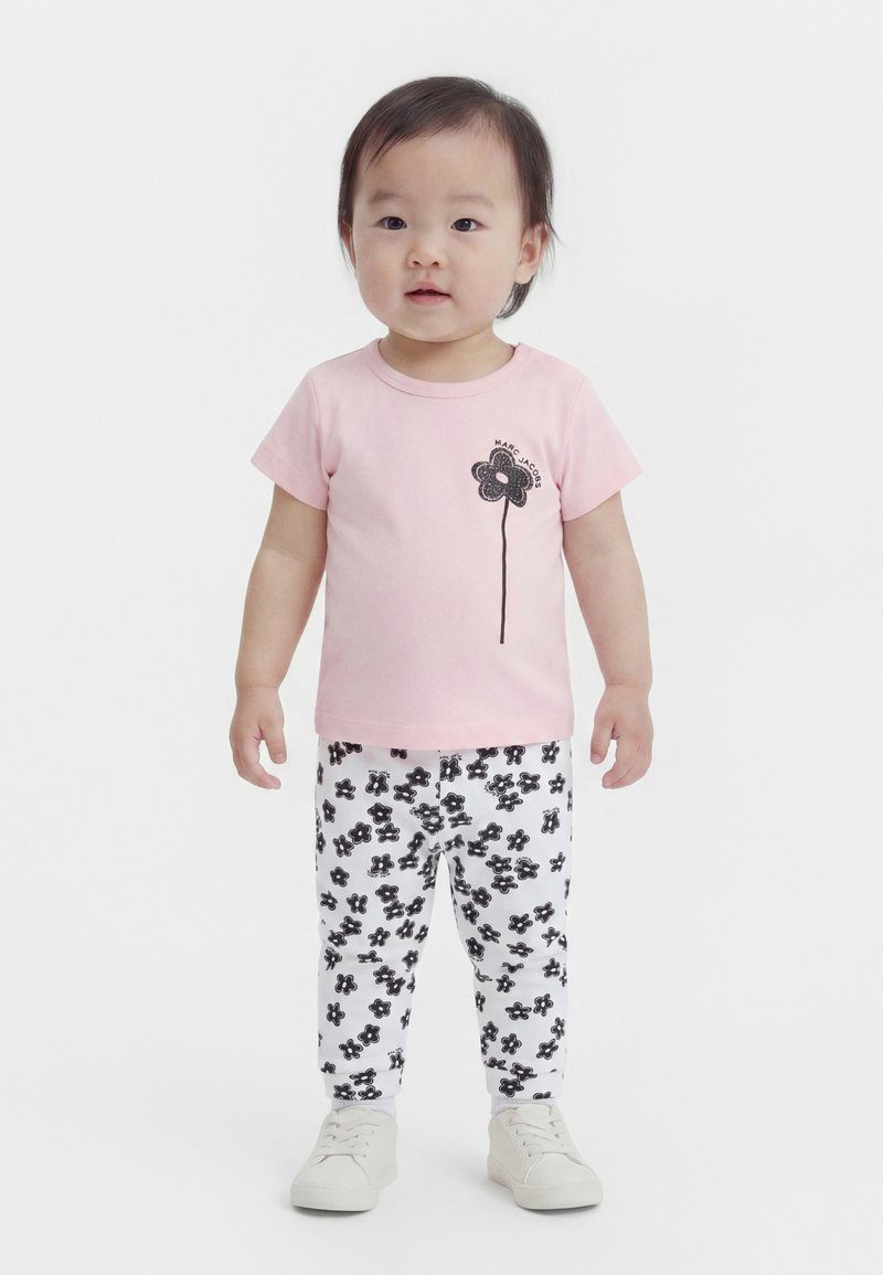 Νήπιο που φοράει ροζ μπλούζα με μαύρο ανθισμένο γραφικό, λευκό παντελόνι με μαύρο ανθισμένο μοτίβο και λευκά αθλητικά παπούτσια, στέκεται και κοιτάζει μπροστά.