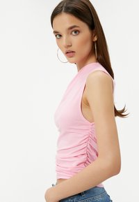 Top sin mangas de color rosa claro con detalles fruncidos en los costados, hecho de material suave; combinado con jeans de mezclilla azules, que realzan un diseño ajustado.