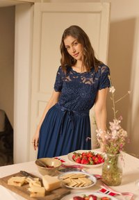 Young woman in a navy blue lace dress stands by a table with cheese, crackers, strawberries, and a vase of flowers in a cozy room.