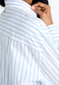 Světle modrá a bílá pruhovaná košile s jemnou texturou, která má límec a mírný záhyb na zadní straně.