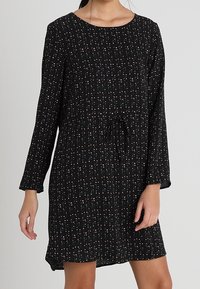 Nainen, jolla on päällään musta pitkähiha dressi, jossa on pieniä vaaleanpunaisia ja valkoisia pilkkuja sekä kiristysnauha vyötäröllä.