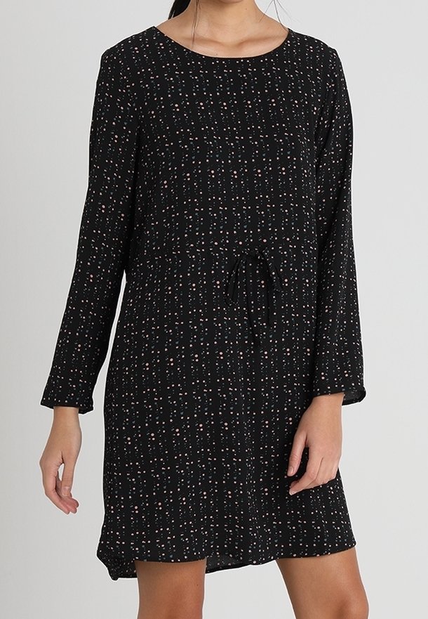 Nainen, jolla on päällään musta pitkähiha dressi, jossa on pieniä vaaleanpunaisia ja valkoisia pilkkuja sekä kiristysnauha vyötäröllä.