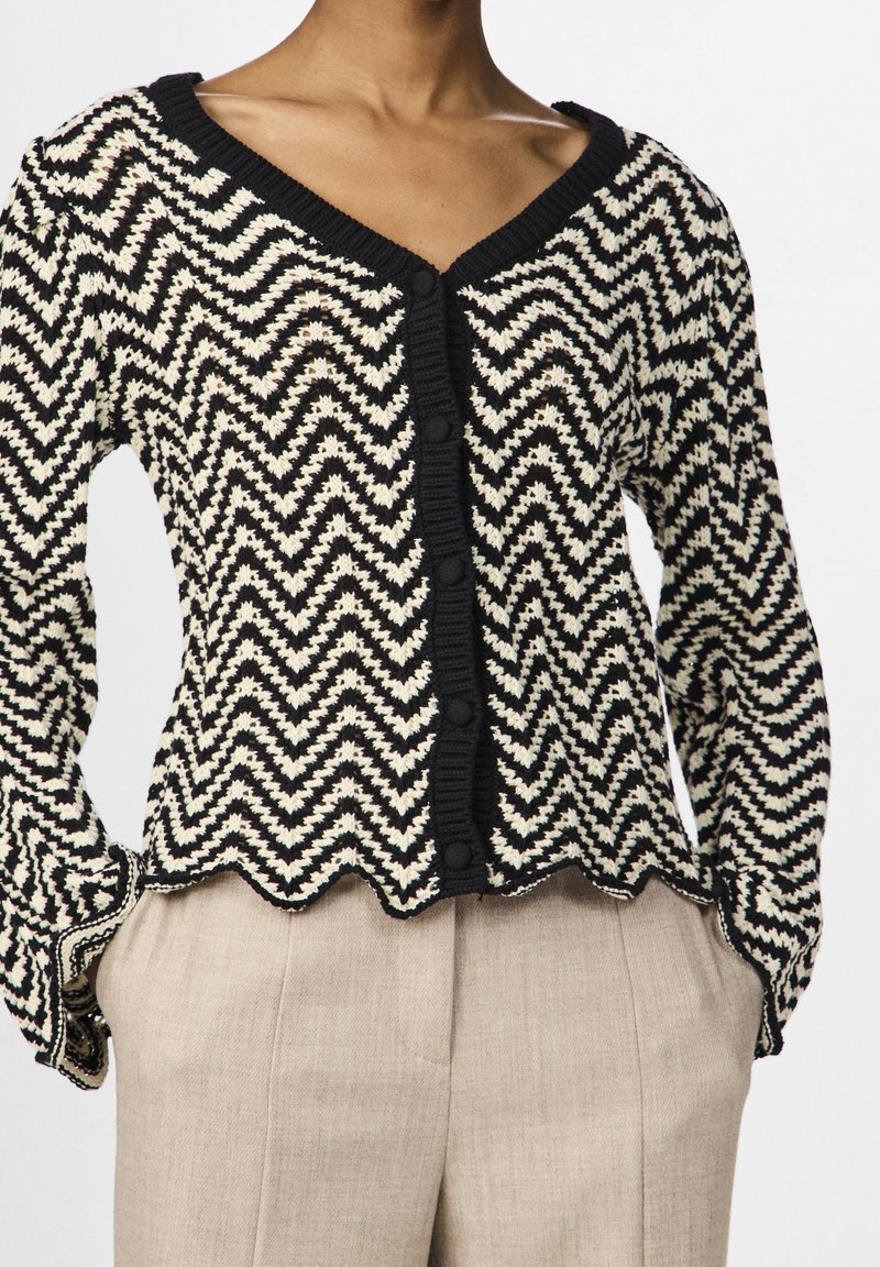 Cardigan tricoté noir et blanc à motif zigzag avec bords festonnés porté sur un pantalon beige taille haute sur une personne debout.