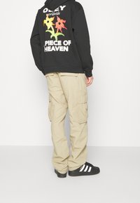 Černá mikina s barevným květinovým vzorem a textem "OBEY worldwide PIECE OF HEAVEN" na zadní straně; kombinováno s khaki cargo kalhotami a černými botami.