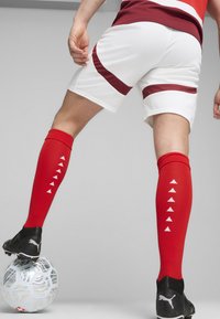 Sarkanas futbola zeķes ar baltiem trīsstūrveida rakstiem, baltas bikses ar sarkaniem svītrām un melnas futbola kurpes uz rakstainas bumbas.