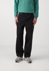 Black denim jeans with a straight-leg cut, featuring a standard five-pocket design and silver button hardware. Worn with a green hoodie and sneakers.
