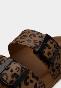 Καφέ σανδάλι με μαύρο λεοπάρ σχέδιο και δύο ρυθμιζόμενες μαύρες αγκράφες σε ιμάντες πάνω από την πέλμα.