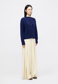 Γυναίκα που στέκεται φορώντας ένα ναυτικό μπλε πλεκτό καλσόν με κουμπιά πάνω από το αντίστοιχο τοπ, κρεμ φαρδιά πλισέ παντελόνια και καφέ παπούτσια.