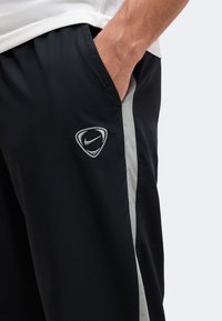 Άτομο που φοράει μαύρη αθλητική βερμούδα Nike με λευκές πλαϊνές ρίγες και το χέρι στην τσέπη, εμφανιζόμενο από τη μέση μέχρι το μέσο του μηρού.