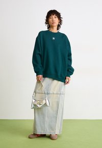 Personne portant un sweat à capuche Adidas teal et une longue jupe en denim avec des rayures blanches, tenant un petit sac à main en argent, se tenant sur un sol vert.