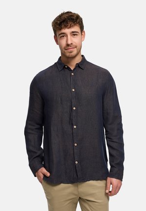 Ung mand med kort krøllet hår iført en mørk knappet skjorte og beige bukser, stående med den ene hånd i lommen foran en ensfarvet baggrund.