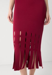 Czerwona dzianinowa sukienka z pionowymi wycięciami wzdłuż dołu. Materiał jest prążkowany i dopasowany, z lekko rozszerzającym się dolnym brzegiem.