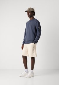 Navy sweatshirt with a black logo patch, cream shorts with "Calvin Klein" printed, white sneakers, and beige cap. Simple, casual outfit.