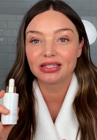 Bouteille blanche avec un compte-gouttes, accents dorés et design minimaliste. L'arrière-plan présente des carreaux gris et une lumière circulaire.