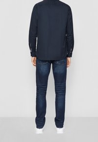 Man i mörkblå långärmad skjorta, mörkblå jeans och vita sneakers, stående mot en ljusgrå vägg.