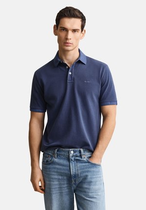 Młody mężczyzna w granatowej krótkiej koszulce polo i jasnoniebieskich jeansach, stojący z jedną ręką w kieszeni na jednolitym tle.