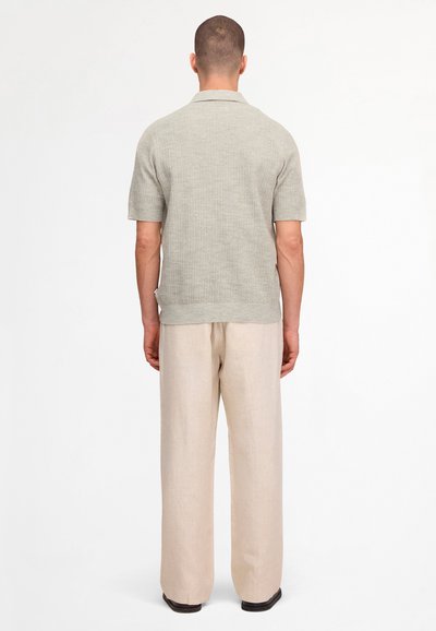 Uomo di spalle, indossa una maglia di maglia grigio chiaro a maniche corte e pantaloni beige larghi con scarpe nere, in piedi su sfondo bianco.