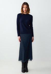 Marineblauwe trui met lange mouwen gecombineerd met een doorschijnende, donkerblauwe rok met een subtiel patroon, gedragen met knielange zwarte leren laarzen.