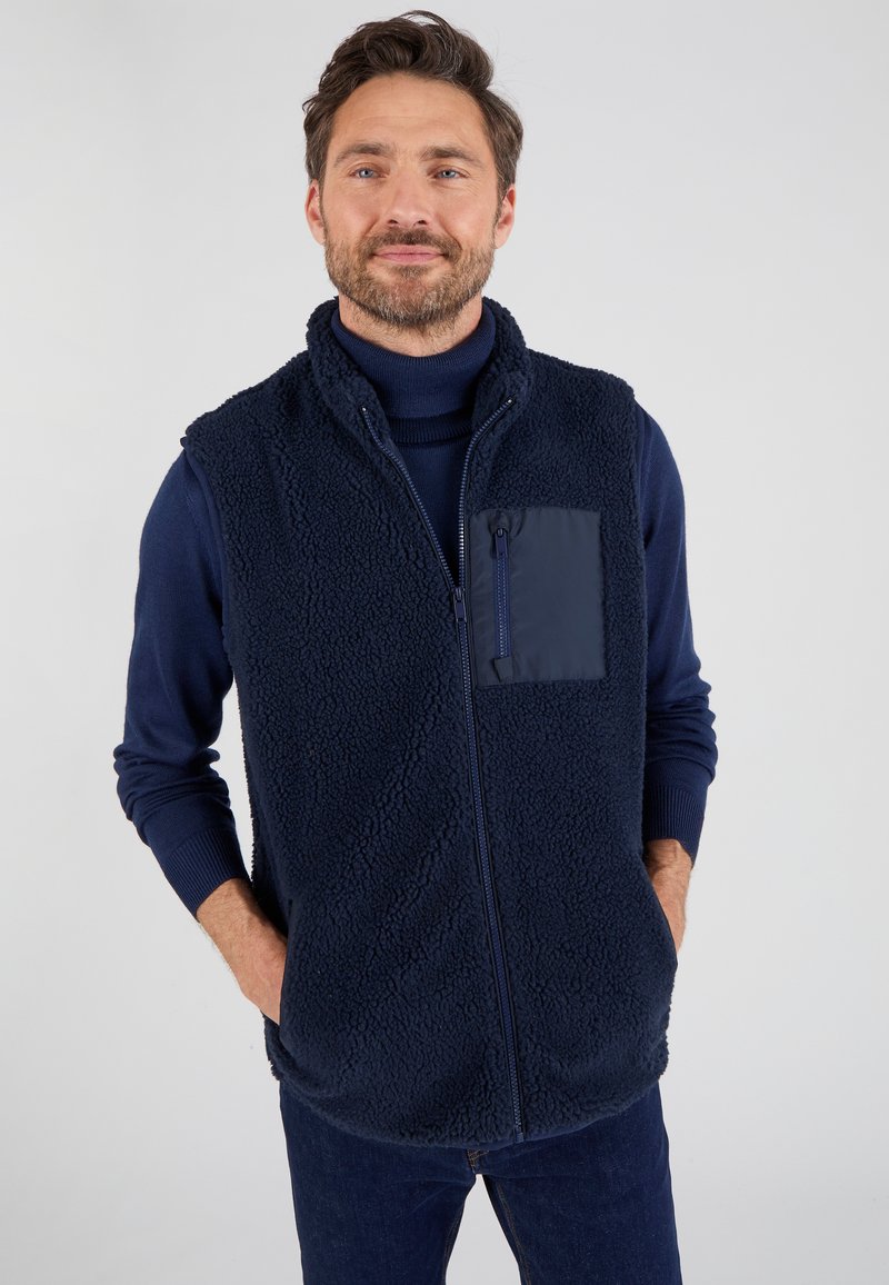 Marineblaue Fleece-Weste mit einer weichen, strukturierten Oberfläche, ausgestattet mit einer Reißverschlusstasche auf der Brust und einem hohen Kragen für zusätzlichen Wärme.