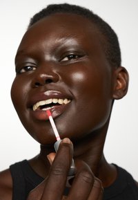 Błyszczyk w przezroczystej tubce z różowym aplikatorem, nałożony na ciemne usta. Modelka ma ciemną skórę i krótkie, teksturowane włosy, nosi czarną bluzkę.