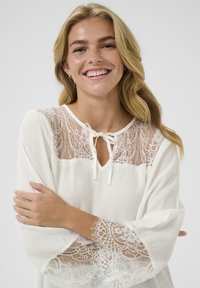 Weißes Blusenshirt mit Spitze am Yoke, Bindekragen und ausgestellten Ärmeln, die mit filigranen Spitzenakzenten an den Bündchen versehen sind. Weicher, leichter Stoff.