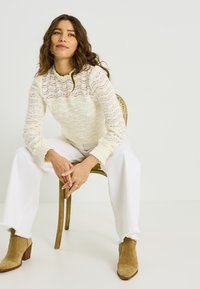 Suéter de punto blanco con encaje y escote con borde festoneado, combinado con pantalones blancos de pierna ancha y botines de gamuza color beige. Cabello rizado y postura sentada.