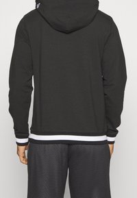 Haut à capuche noir avec une capuche, présentant une taille rayée noire et blanche. Le matériau semble doux. Porté avec un short de sport noir texturé.