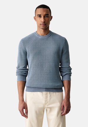 Uomo che indossa un maglione a coste azzurro chiaro con le maniche rimboccate e pantaloni beige, in piedi davanti a uno sfondo chiaro e semplice.