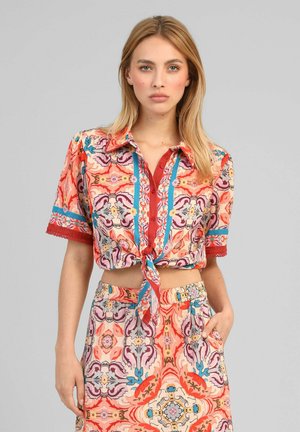Femme portant une chemise à manches courtes à motifs colorés, boutonnée et nouée à la taille, et un pantalon taille haute assorti, debout avec une main dans la poche.