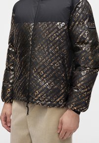 Μαύρο και χρυσό jacket EA7 Emporio Armani με φερμουάρ και σχέδιο, φορεμένο από ένα άτομο με μπεζ παντελόνια, που δείχνει μόνο το σώμα και τα χέρια.