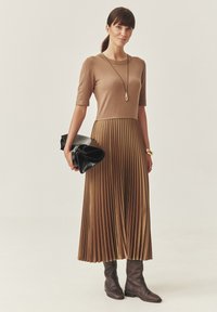 Vestido marrón con pliegues, de corte ajustado en la parte superior y mangas cortas, combinado con botas marrones hasta la rodilla y un bolso de mano negro. Textura suave, diseño elegante.